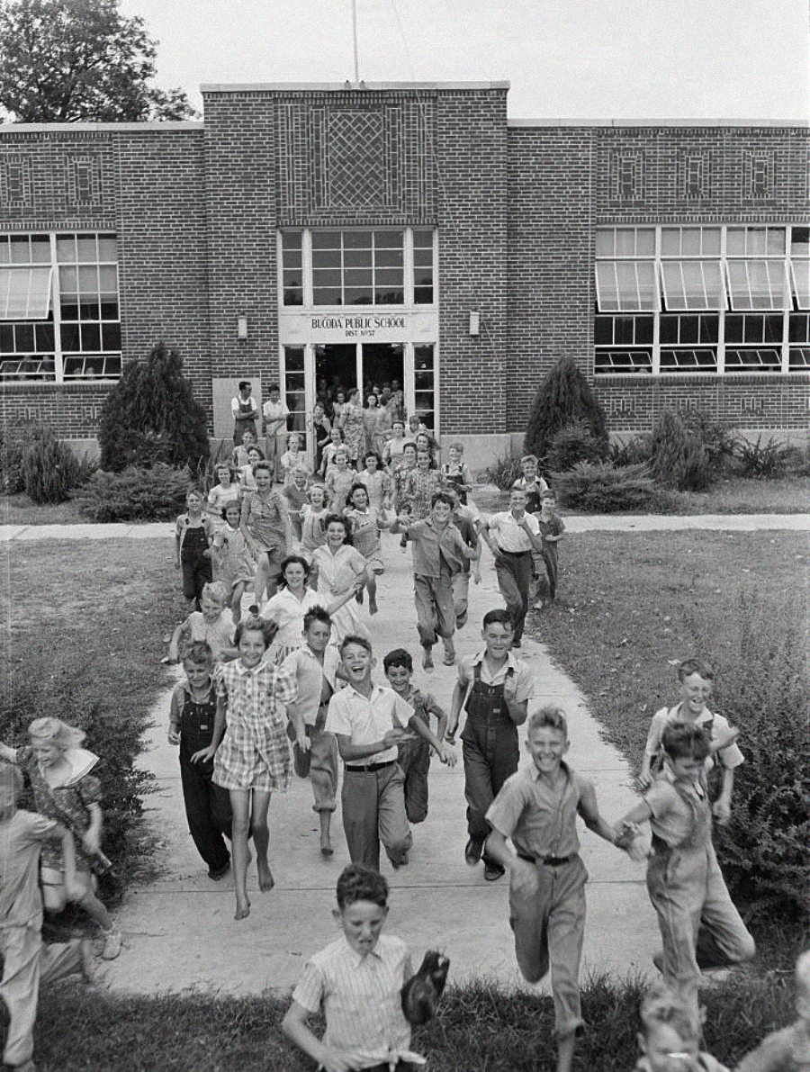 A large group of children, smiling and excited, run out of the front entrance of a brick school building labeled "Buena Public School" on a sunny day. Shrubs and grassy areas line the walkway.