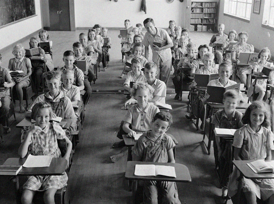 A black-and-white photo of a classroom filled with young students sitting at desks. Most of the children are smiling and facing the camera, while a teacher stands at the back, also looking at the camera.
