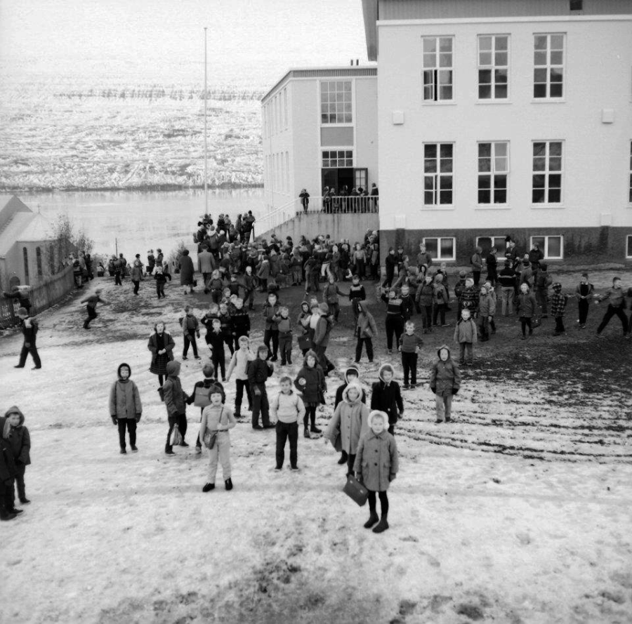 A large group of children bundled in winter clothes stand and play on a snowy, slushy ground outside a white building, possibly a school, with more children visible in the background near a waterfront.