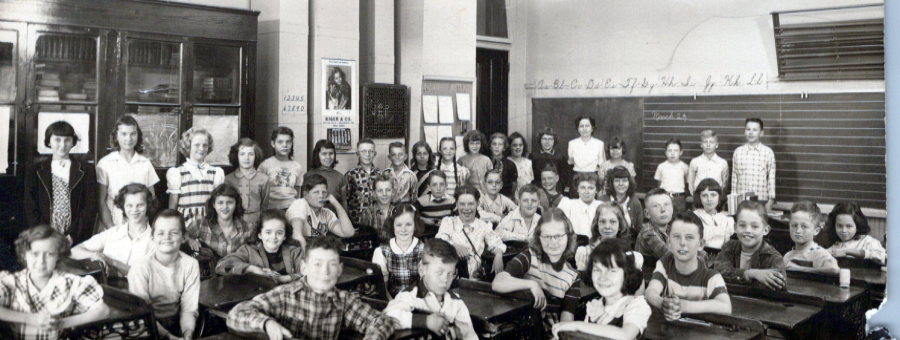 Black-and-white photo of a large classroom filled with young students sitting at desks and standing, with two teachers at the back. The room has a chalkboard, posters, and shelves. The children are smiling and facing the camera.