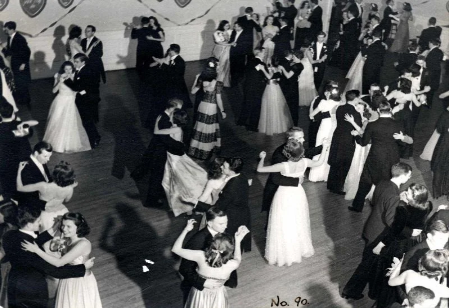 Black and white photo of couples in formal attire dancing in a large ballroom. Women wear long gowns and men wear suits or tuxedos. The setting appears to be a formal event or prom from the mid-20th century.