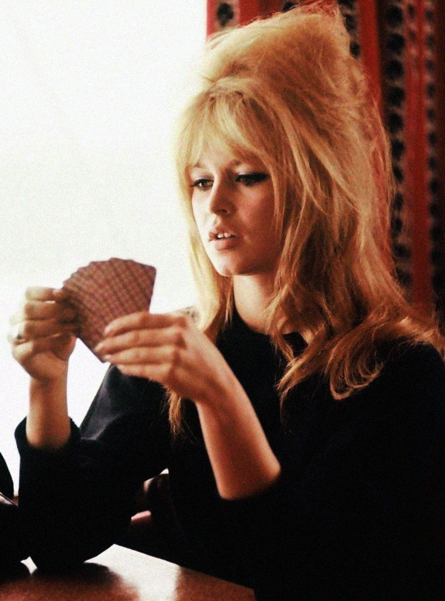 A woman with voluminous blonde hair and a black top is sitting at a table, holding and looking at a hand of playing cards. The background is softly lit with patterned red and black curtains.