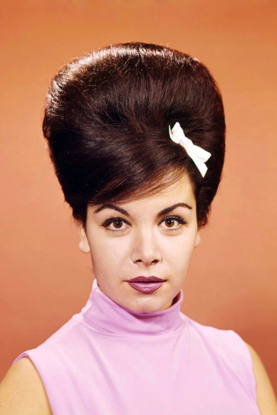 Woman with dark, voluminous beehive hairstyle adorned with a white bow, wearing a sleeveless light pink turtleneck top, poses against a plain peach background.