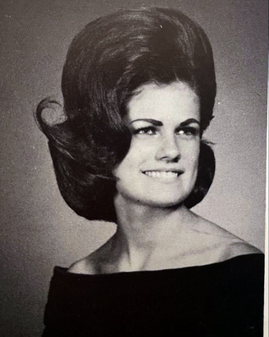 Black and white portrait of a woman with a large, voluminous beehive hairstyle, wearing an off-the-shoulder top and smiling, looking slightly to the side.