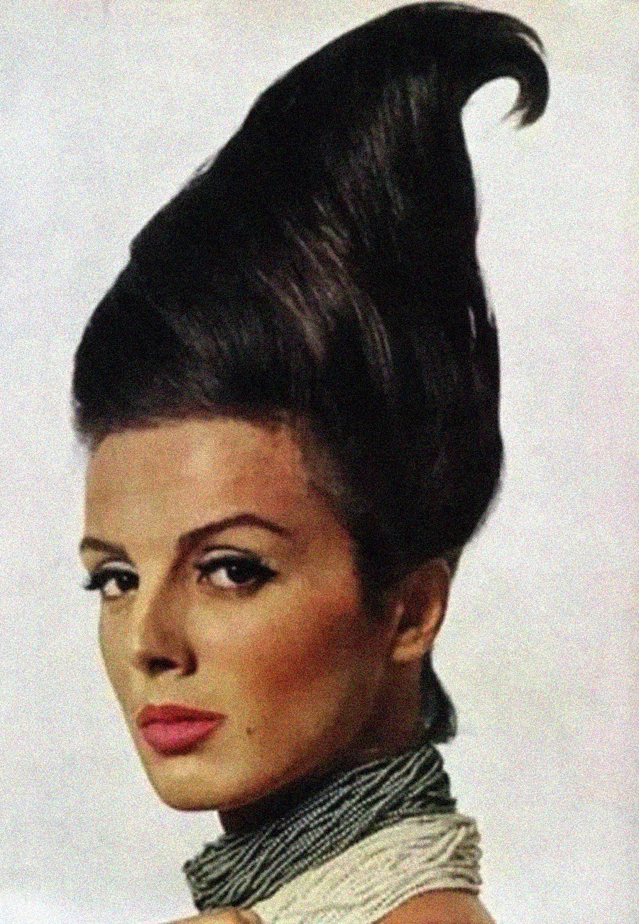 A woman with dramatic makeup poses with her dark hair styled in a tall, sculpted, upward swirl, resembling a conical or pointed shape. She wears a textured, light-colored scarf around her neck.