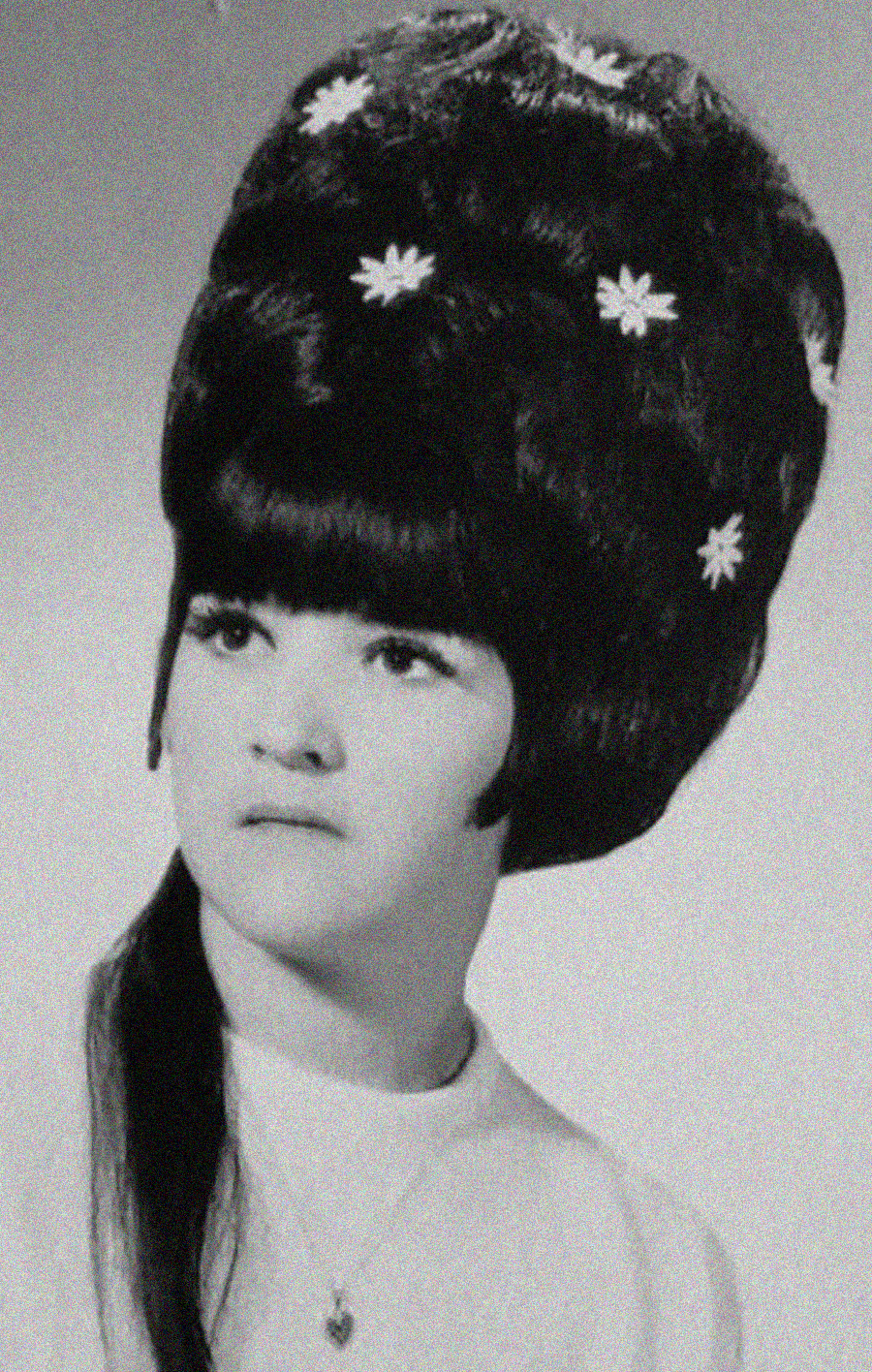 Black and white portrait of a woman with a large beehive hairstyle decorated with small white flowers, wearing a light-colored top and a necklace, looking slightly to the side.
