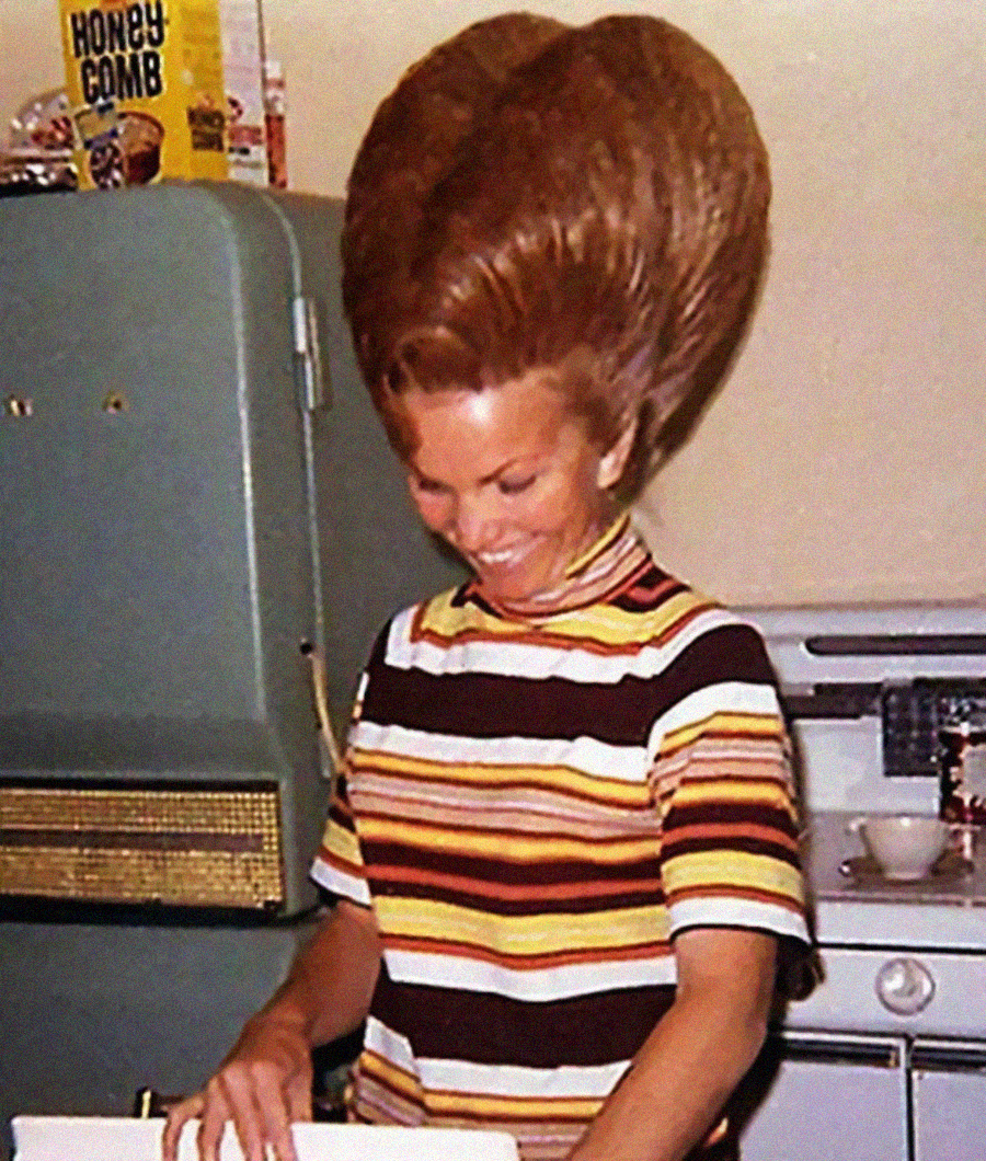 A woman in a retro kitchen smiles while wearing a striped shirt; her hair is styled in an exaggeratedly large beehive, towering above her head. A box of Honey Comb cereal sits on top of a green refrigerator.