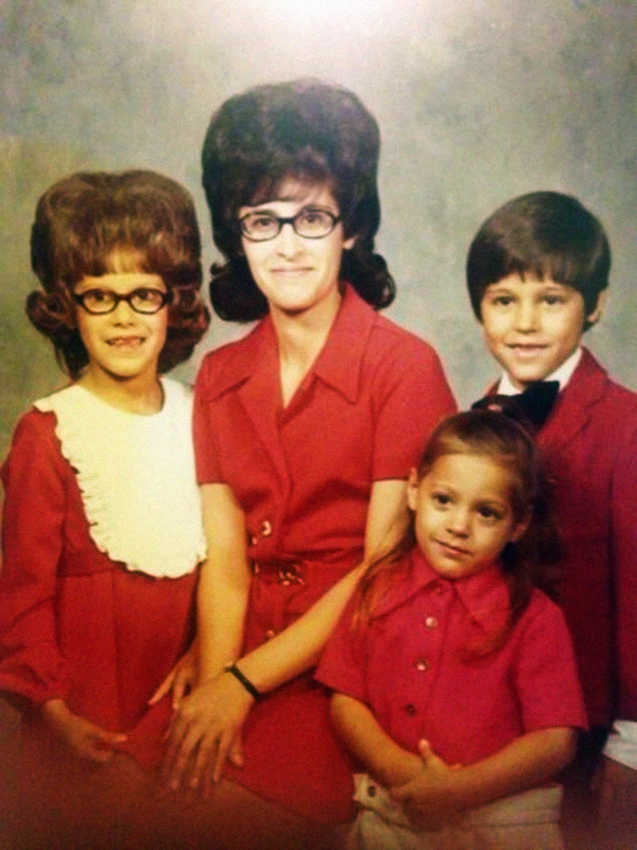 A vintage studio portrait of a woman and three children. The woman and older girl wear red dresses and glasses with large hairstyles. The boy wears a red jacket and bowtie, and a young girl stands in front in a bright pink shirt.