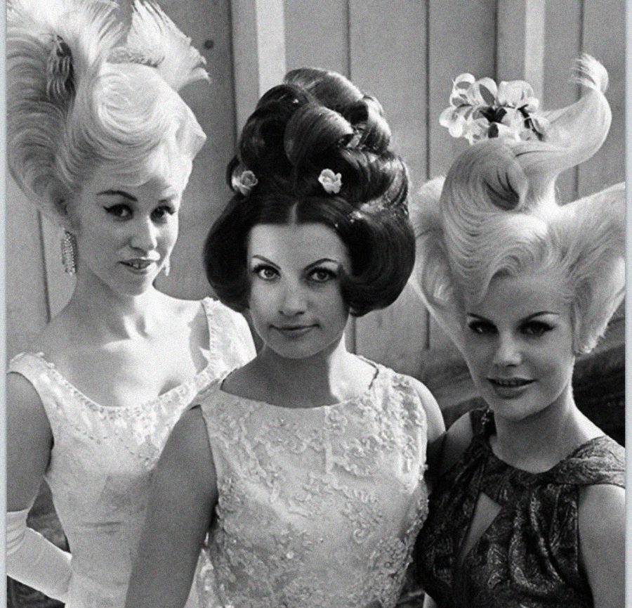 Three women pose together, each wearing extravagant, sculpted 1960s-era hairstyles. Two have high, swooping hairdos, and one has curls and decorative flowers. All wear patterned dresses and smile slightly for the camera.