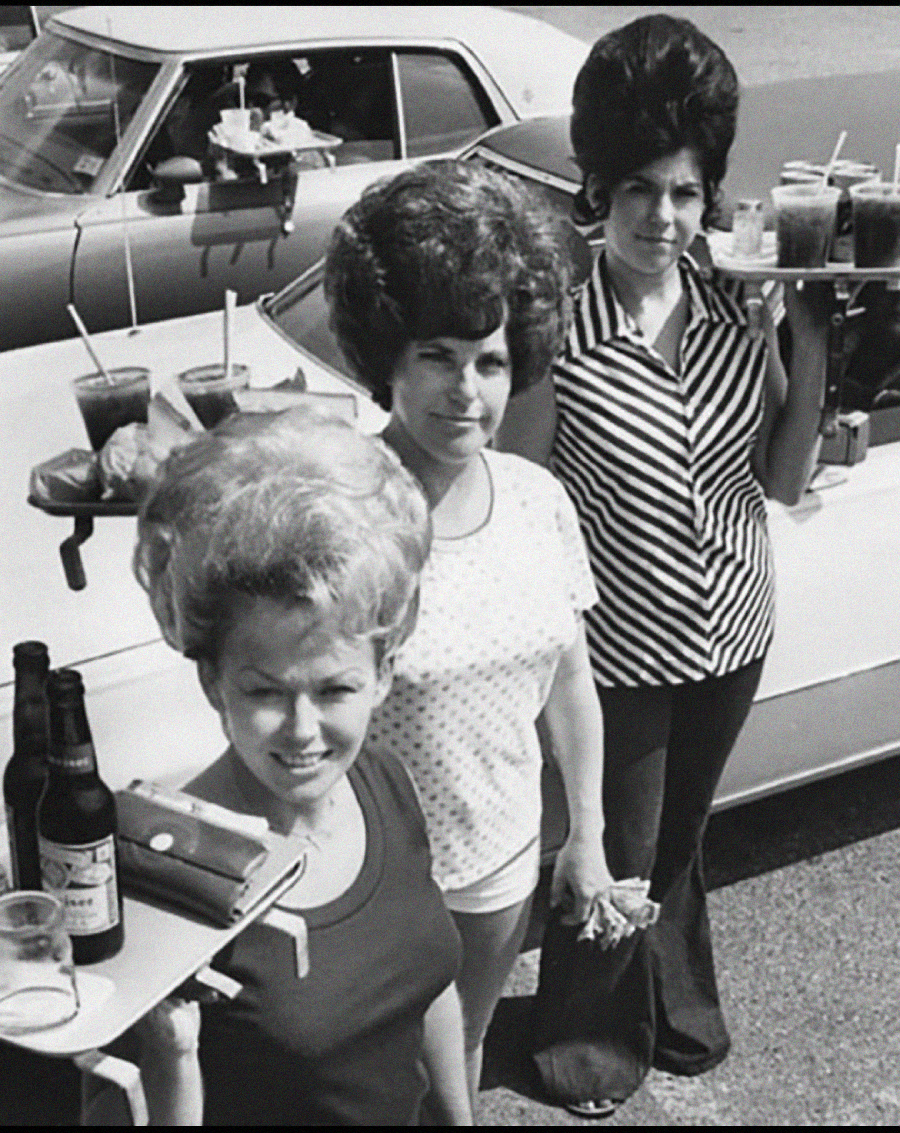 Three women with beehive hairstyles stand by cars, holding trays with drinks and food, serving as carhops in a retro outdoor drive-in setting. A classic car with trays attached to the window is in the background.