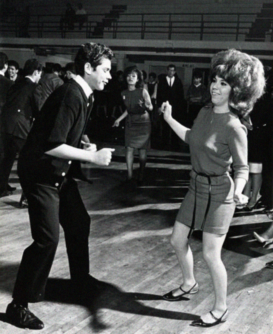 A man and woman dance energetically on a wooden floor in a crowded hall. The woman has a beehive hairstyle and wears a short dress, while the man wears a dark jacket and pants. People in the background also appear to be dancing.
