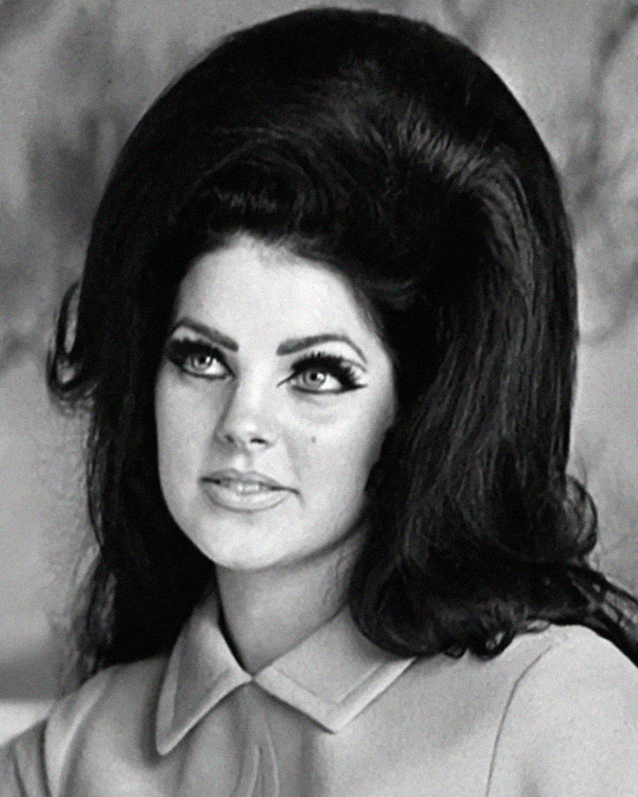 Black and white photo of a woman with voluminous dark hair styled in a beehive, wearing winged eyeliner and a collared top, looking slightly to the side with a neutral expression.