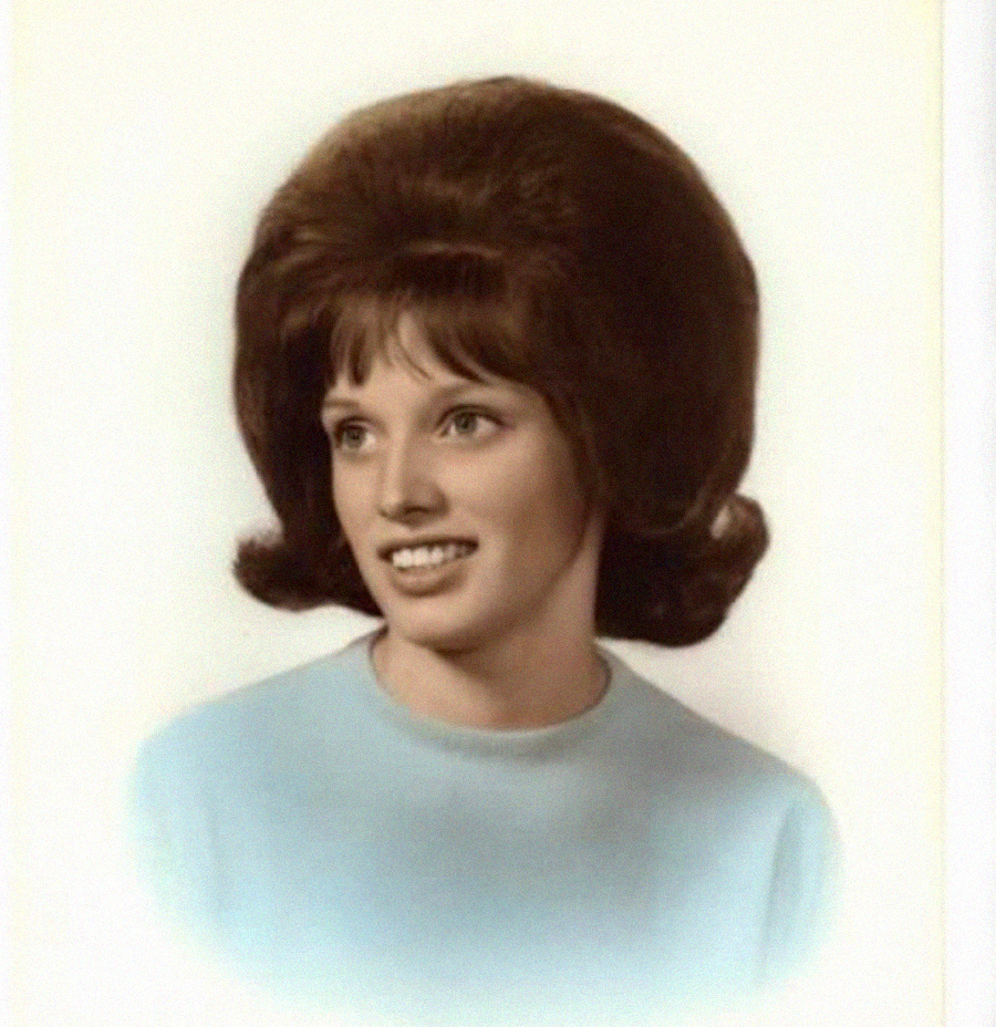 A young woman with light skin, wearing a light blue top, smiles while looking slightly to the side. She has a voluminous brown beehive hairstyle and is photographed against a plain, light background.