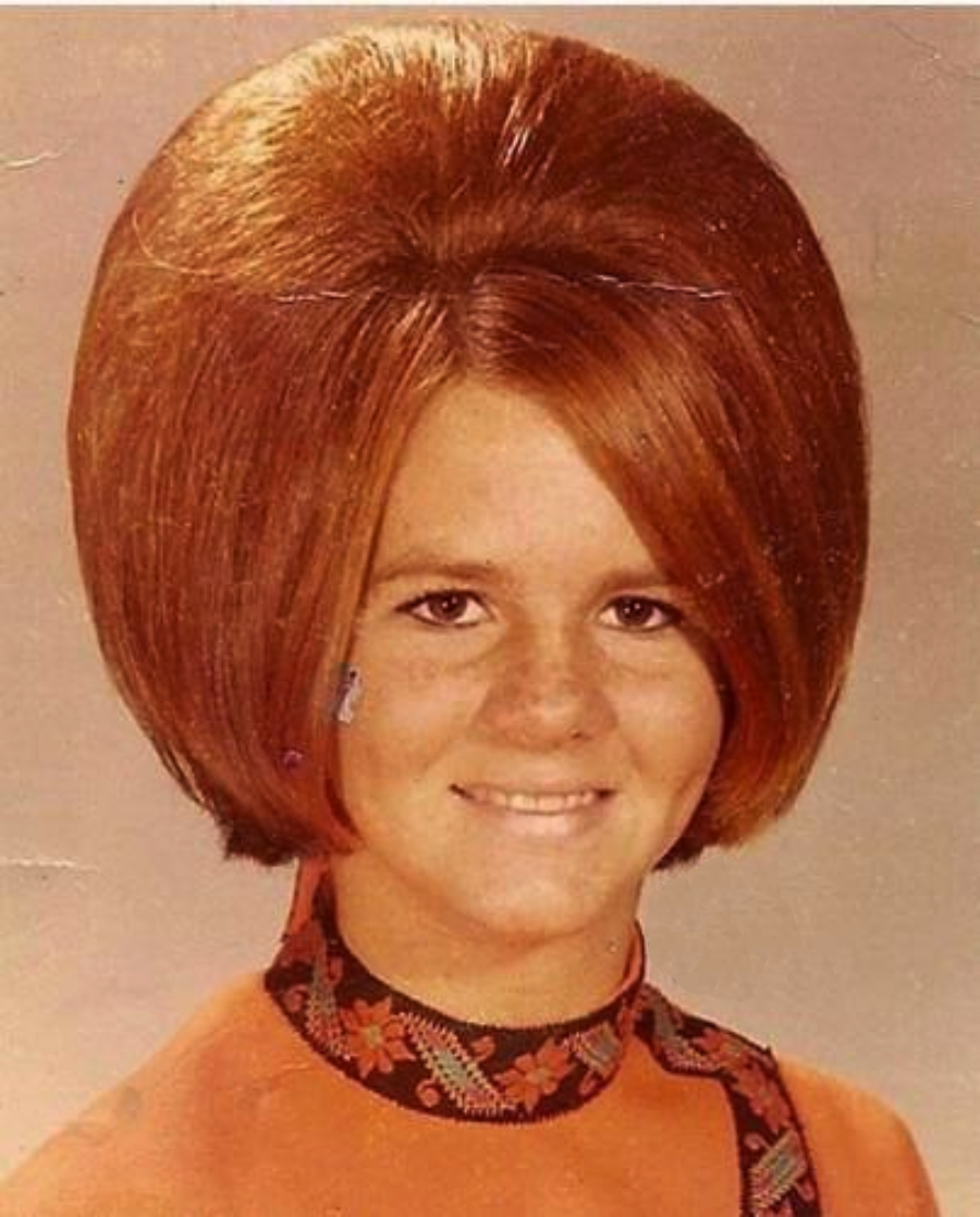 A young woman smiles at the camera, wearing an orange top with a patterned collar and sporting a large, voluminous bouffant hairstyle characteristic of the late 1960s or early 1970s.