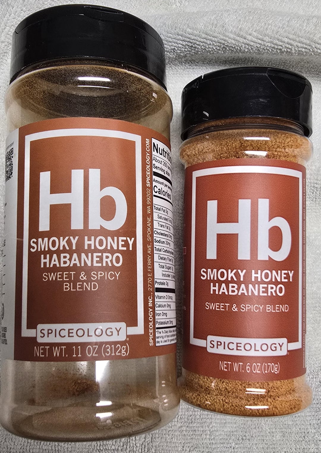 Two spice containers labeled "Hb Smoky Honey Habanero Sweet & Spicy Blend" from Spiceology, one 11 oz (312g) and the other 6 oz (170g), are placed side by side on a light fabric background.