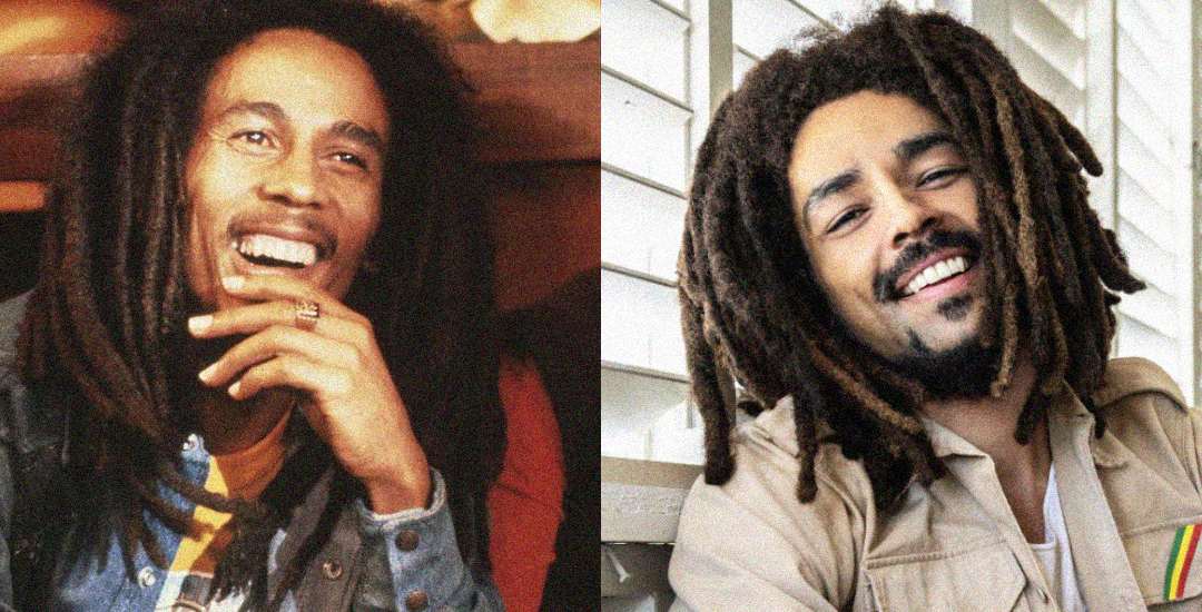 Two men with long dreadlocks and beards are smiling. The man on the left wears a dark jacket and has his hand near his chin. The man on the right wears a light shirt and is sitting in front of a window with white blinds.