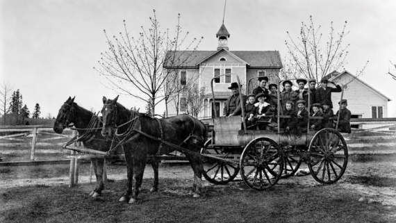A black-and-white photo of children and adults sitting in a horse-drawn wagon in front of a wooden house with a small bell tower, surrounded by leafless trees and a wooden fence.