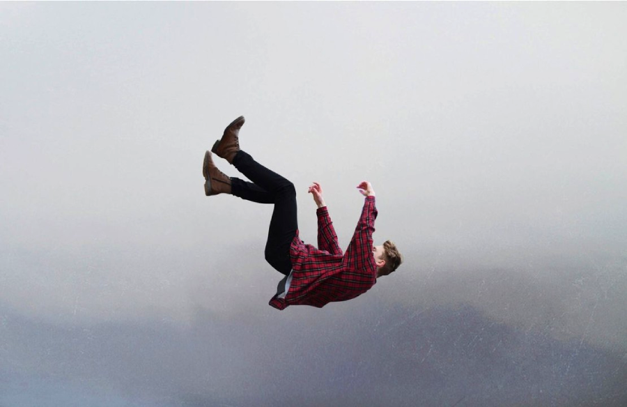 A person wearing a red plaid shirt, black pants, and brown boots is suspended mid-air, falling backwards against a cloudy, grey background.