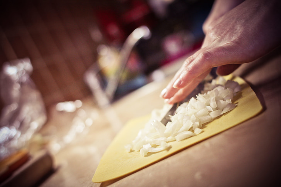 A close-up of a hand chopping white onions on a yellow cutting board in a kitchen, with a knife and blurred background visible.