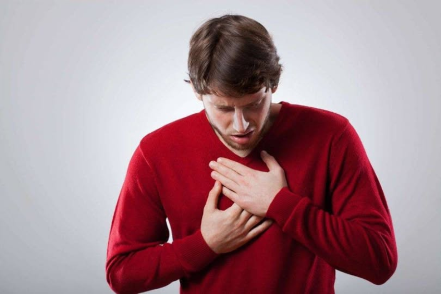 A man in a red sweater holds his chest with both hands and looks down with a concerned expression, possibly indicating chest pain or discomfort, against a plain gray background.
