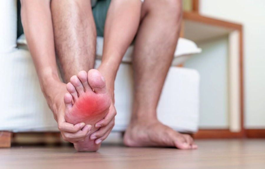 A person sits on a couch holding their foot, with the bottom of the foot showing a red, irritated area, indicating pain or inflammation. The other foot rests flat on the wooden floor.