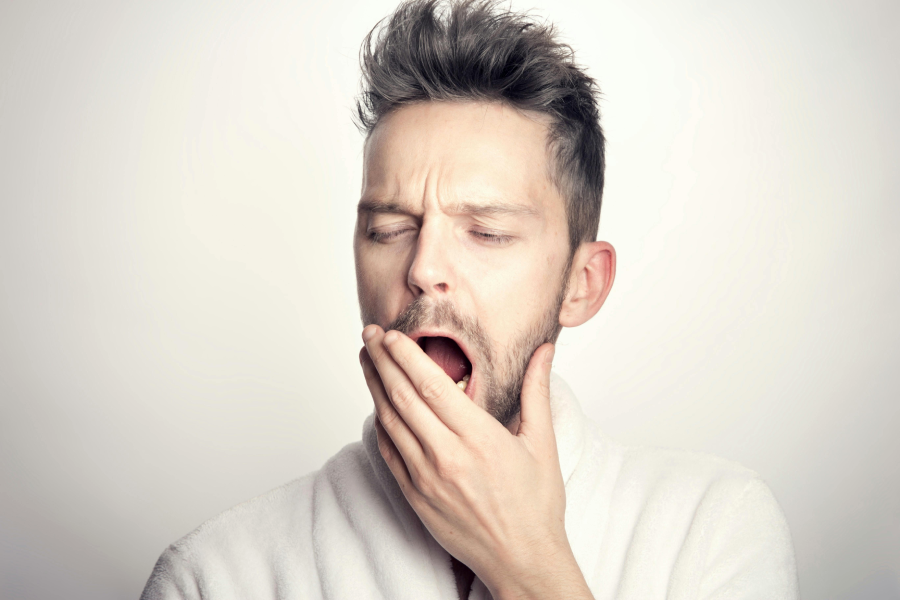 A man wearing a white bathrobe yawns and covers his mouth with one hand, appearing tired or sleepy, against a plain light background.