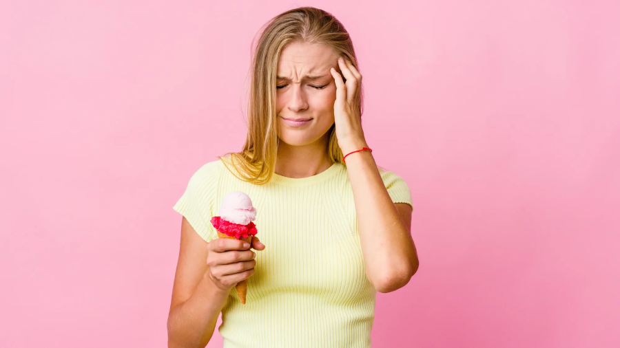 A young woman holding an ice cream cone in one hand and touching her forehead with a pained expression, standing against a pink background.