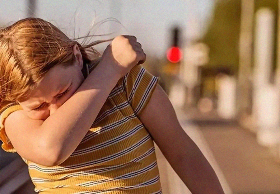 A person in a yellow striped shirt sneezes or coughs into their elbow while standing outdoors near a blurred background of a train station or platform.