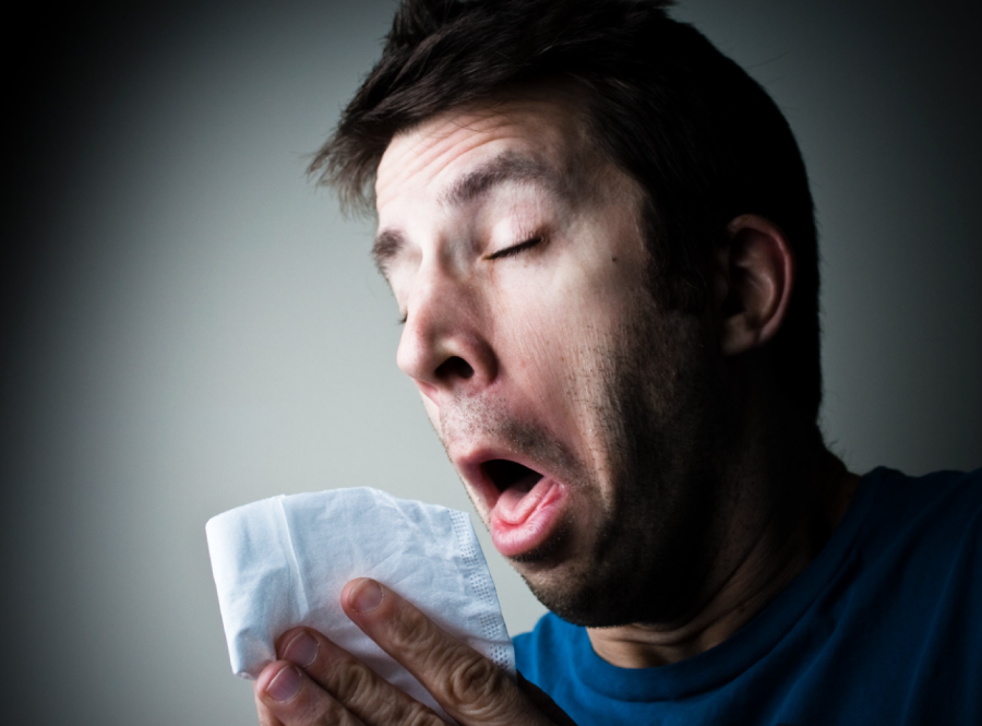 A man sneezes into a white tissue, his eyes closed and mouth open, with a pained expression on his face. The background is plain and out of focus.