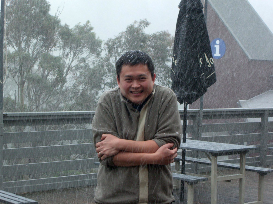 A person stands outside on a deck in heavy rain, shivering with arms crossed for warmth. Trees and outdoor furniture are visible in the background, along with an umbrella and a building.