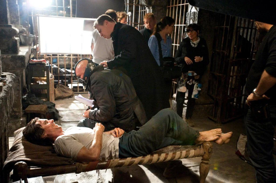 Behind-the-scenes photo from a film set shows a man lying on a cot in a dim, jail-like room as crew members and actors stand or work nearby, preparing for a scene. Film equipment is visible in the background.