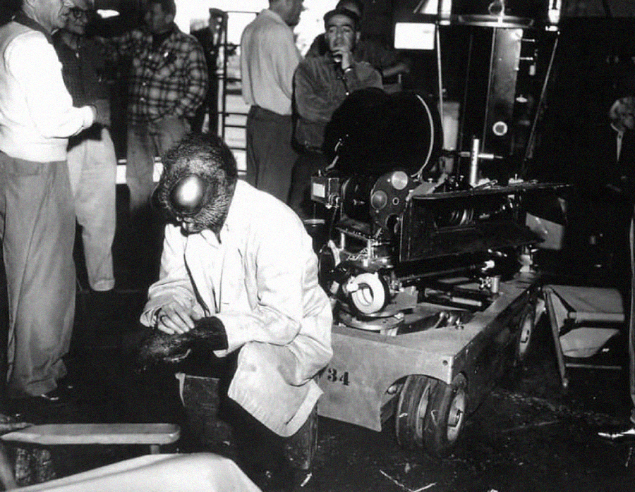 A person in an ape costume kneels on a film set beside camera equipment, while several crew members stand and converse in the background. The image is in black and white.