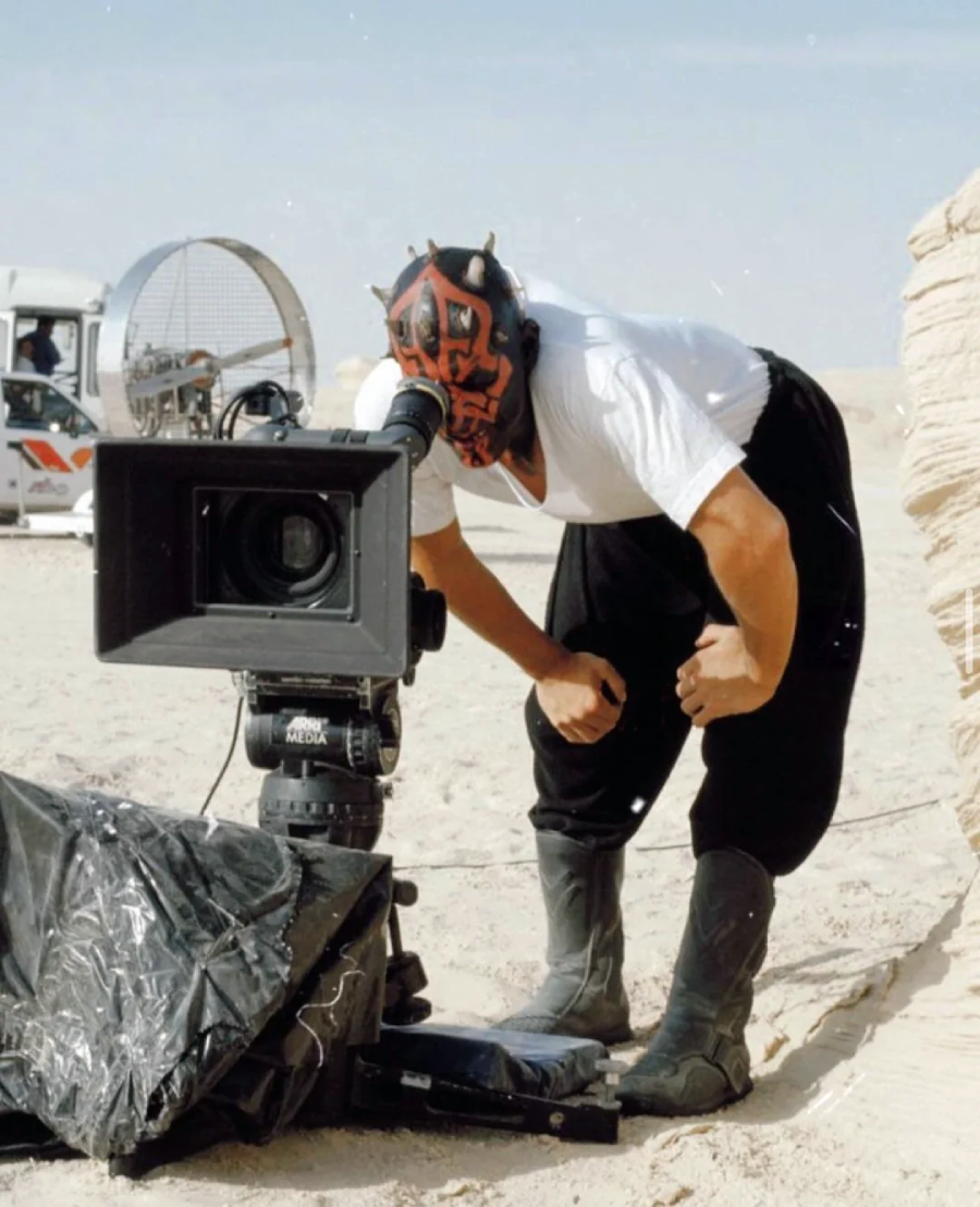 A person wearing a Darth Maul mask and costume looks through a film camera on a desert set, with sand and film equipment visible in the background.