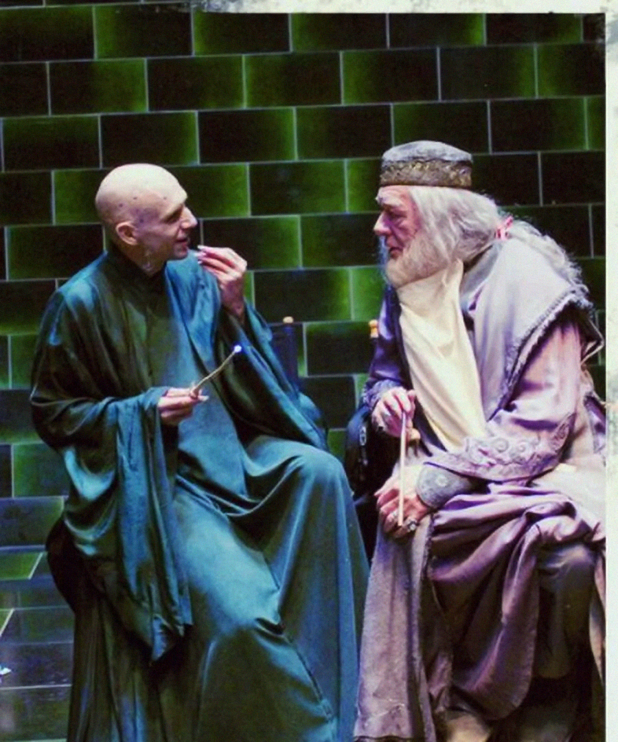 Two men in robes sit facing each other against a green-tiled background, each holding a wand. One is bald and dressed in dark green, the other has long white hair, a beard, and wears a crown and grey robes.