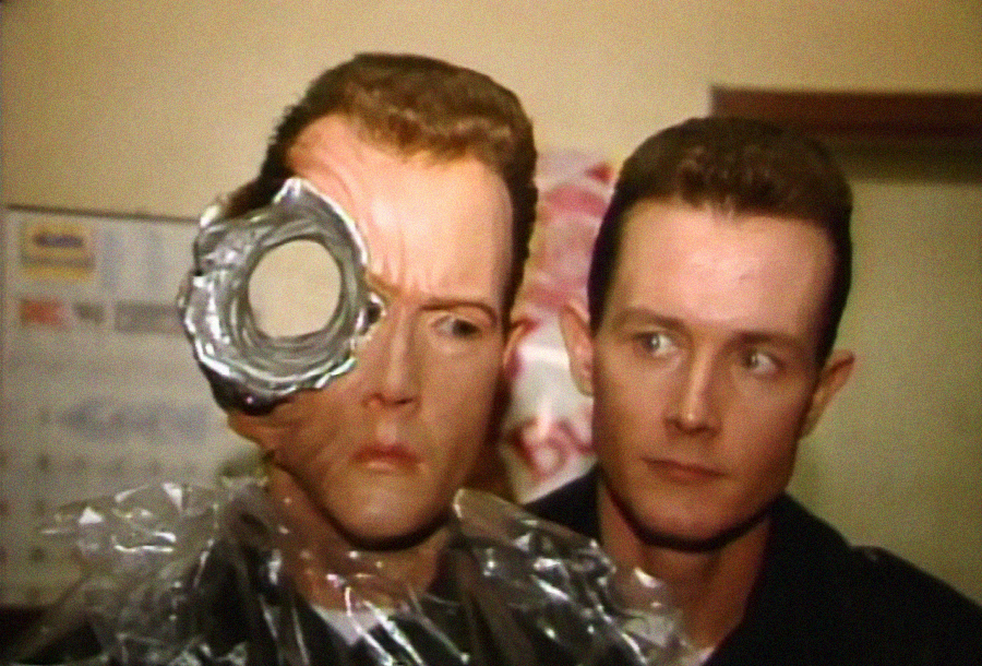 A man stands beside a mannequin head with a large, circular hole through one eye, surrounded by torn metal-like edges, resembling a special effects prop from a sci-fi film.