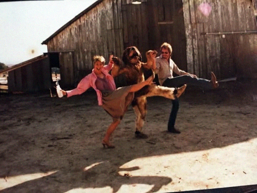 Three people, including one in a hairy, ape-like costume, stand outside a wooden barn, holding hands and kicking their legs high in a playful, coordinated dance pose. It is sunny and the scene looks casual and fun.