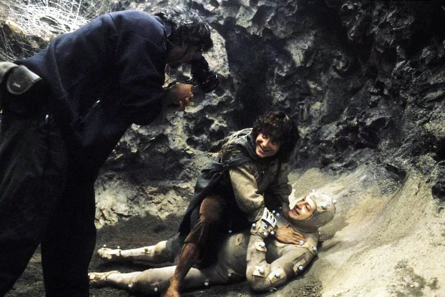 An actor in costume pins down another actor wearing a motion-capture suit on a rocky set, while a cameraman films the scene up close.