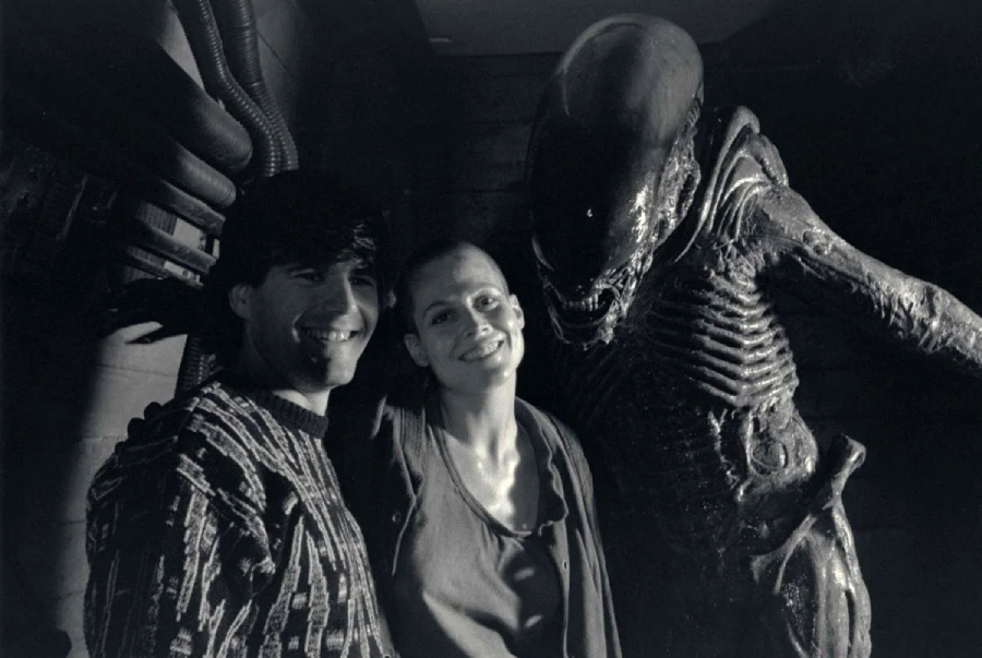 A black-and-white photo shows two people smiling and standing next to a person in an alien creature costume from the Alien film series, posing together in what looks like a film set hallway.