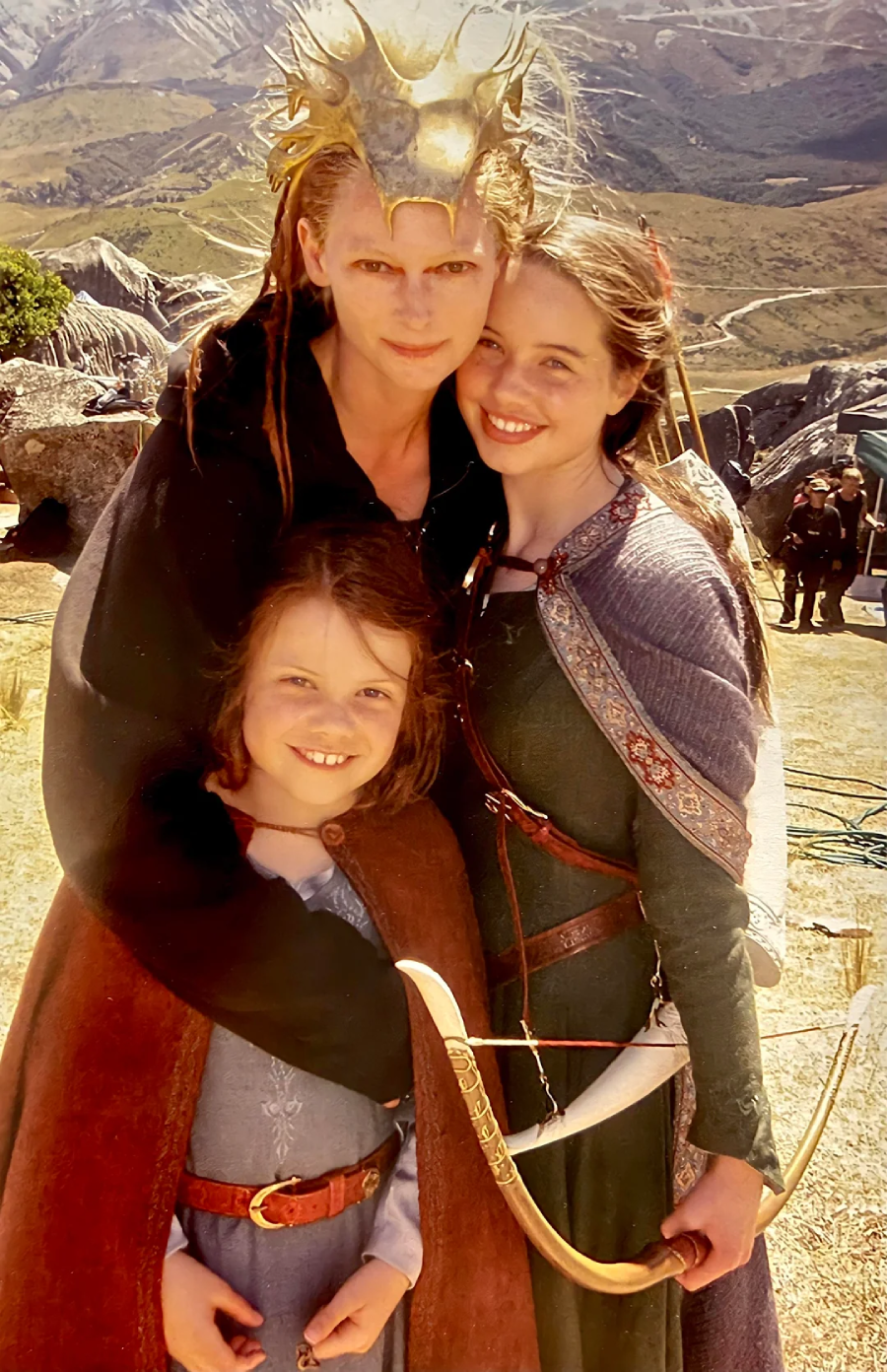 Three people in medieval-style costumes stand outdoors on a grassy hillside. The tallest wears a gold crown and dark robe, hugging the other two, who are dressed in capes. The smallest holds a bow. Mountains are visible in the background.