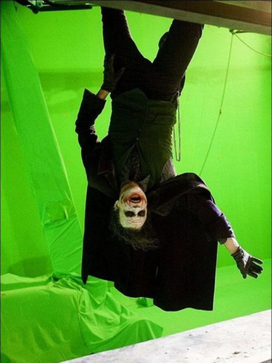 A person dressed as the Joker hangs upside down from a metal bar, with a bright green screen background behind them, likely on a film set.