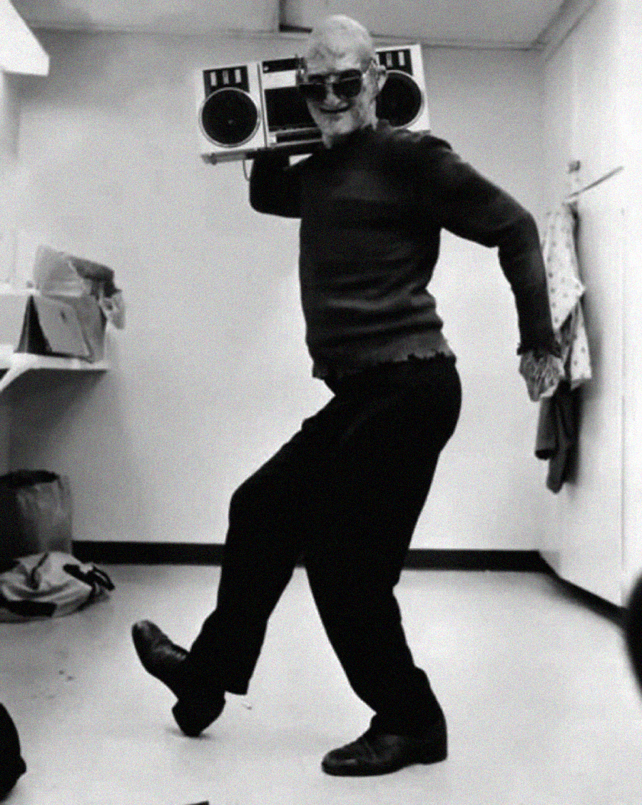 An older man wearing sunglasses strikes a playful dance pose while holding a large boombox on his shoulder in what appears to be a dressing room.