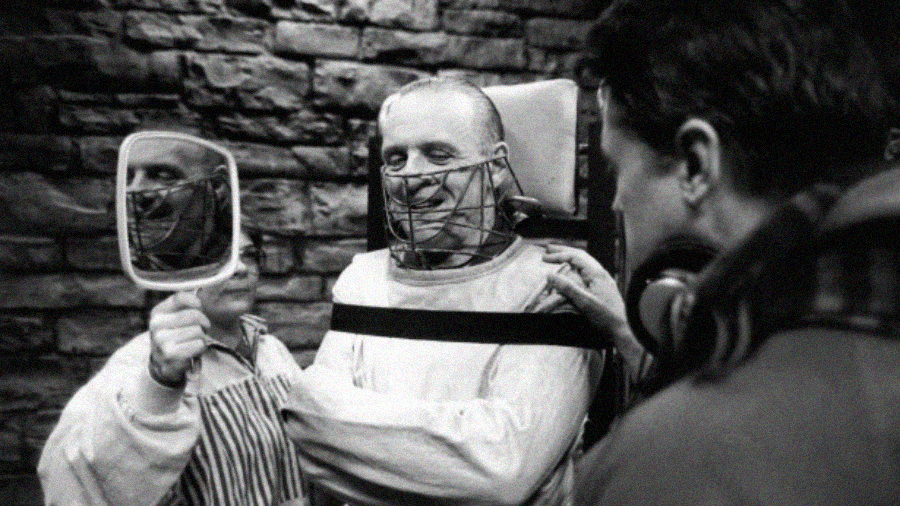 A man in a straitjacket and face mask smiles while looking into a handheld mirror. Another person holds the mirror, and a third person stands nearby. They are set against a stone wall in a black-and-white photo.