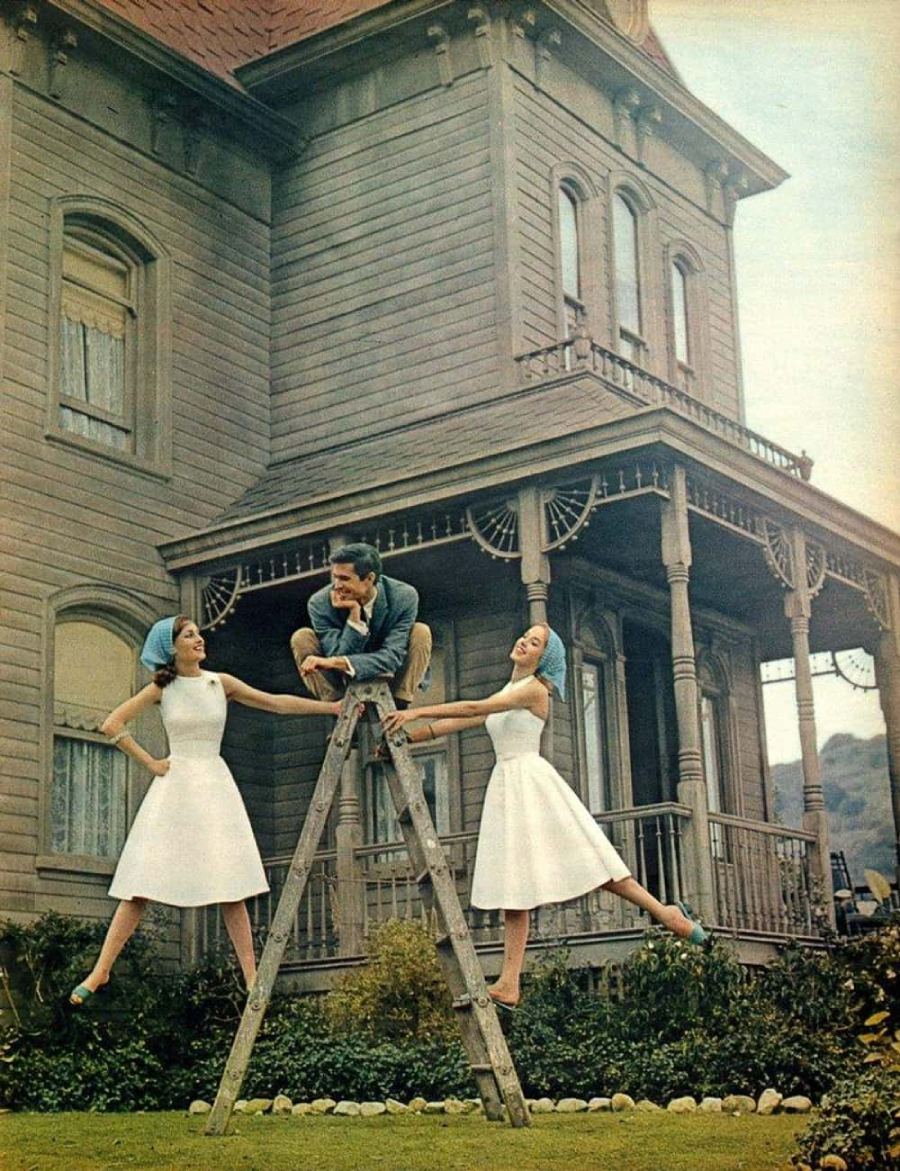 Two women in white dresses and blue headscarves pose on either side of a wooden ladder, each holding a side rail, while a man sits atop the ladder, in front of a large Victorian-style house.
