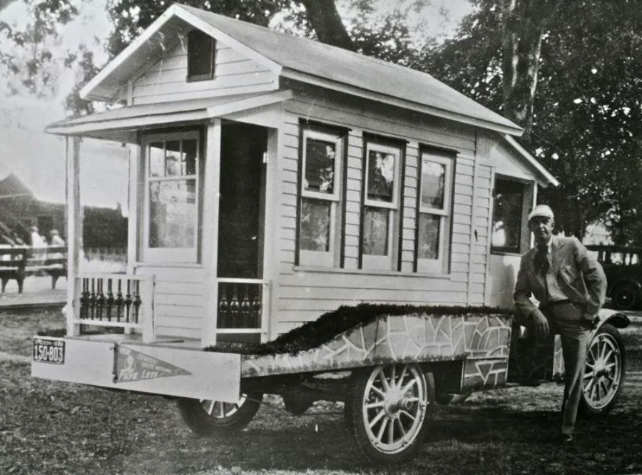 A black and white photo shows a man in a suit and cap standing beside a small house built on a wheeled trailer, resembling a tiny house on wheels, with bottles on its porch and decorative trim.