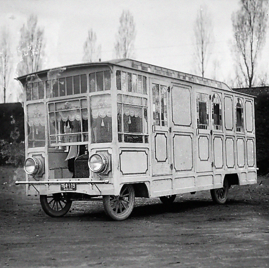 A vintage black-and-white photo of a boxy, early 20th-century vehicle with large glass windows, curtains, and ornate paneling, resembling a small house on wheels, parked outdoors with trees in the background.