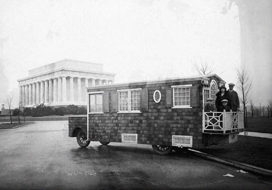 A vintage photo of a shingled, house-like motorhome parked near the Lincoln Memorial. Three people stand on its small porch, and leafless trees dot the background. The image has a historic, black-and-white tone.