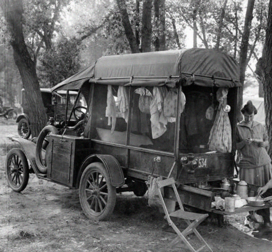 Black and white photo of an old-fashioned camper van with fabric draped in the windows and a small outdoor kitchen setup; a woman prepares food beside the vehicle, surrounded by trees.