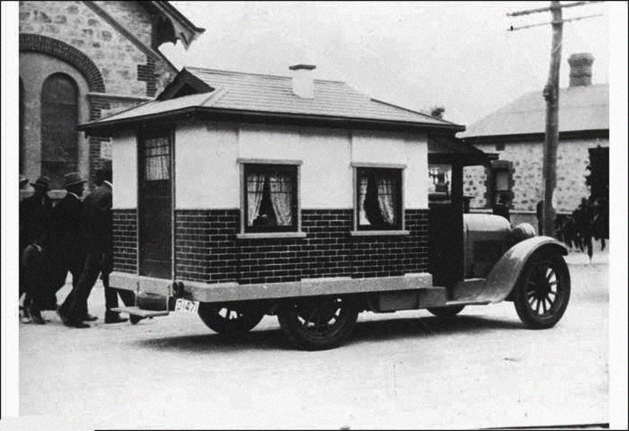 A vintage black-and-white photo shows an old car modified with a small, house-like structure on its bed, complete with brick-pattern siding, windows, and curtains. Several people stand nearby on a street.