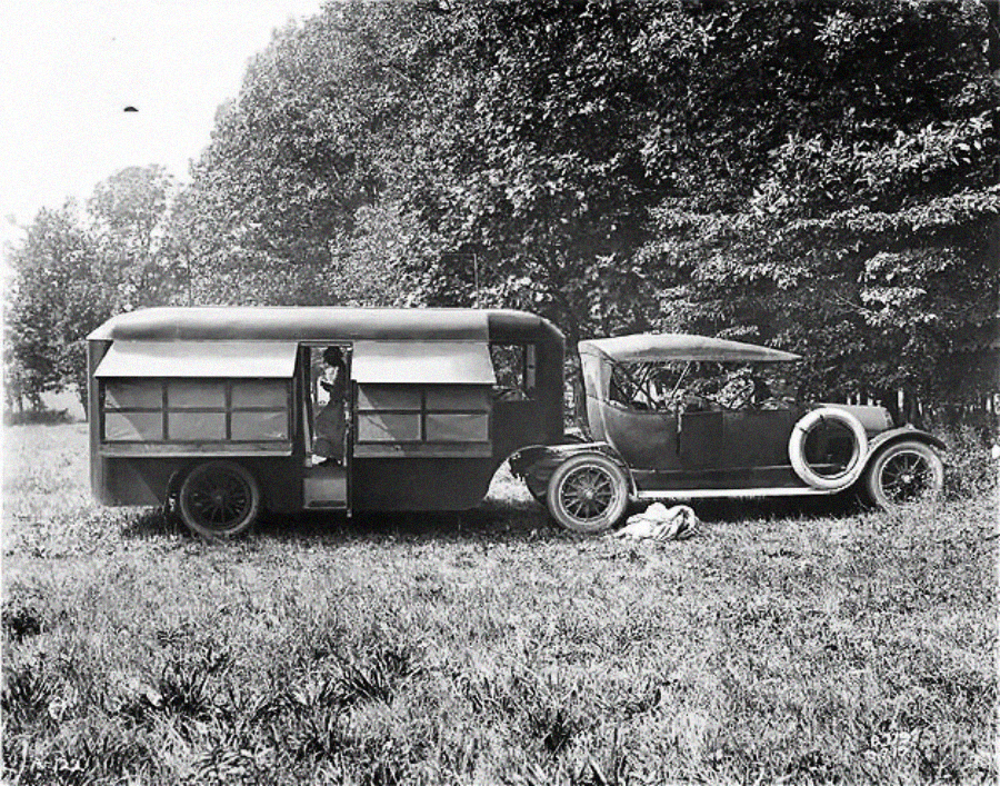 A vintage black-and-white photo shows an old car towing a covered camping trailer in a grassy field, surrounded by trees. A person stands in the trailer doorway, and a dog lies on the ground nearby.