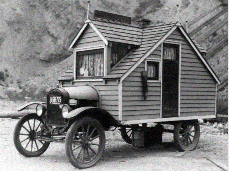 A vintage black-and-white photo shows an old car with a small wooden house structure built on its back, complete with windows, a door, and curtains, resembling an early motorhome or RV.